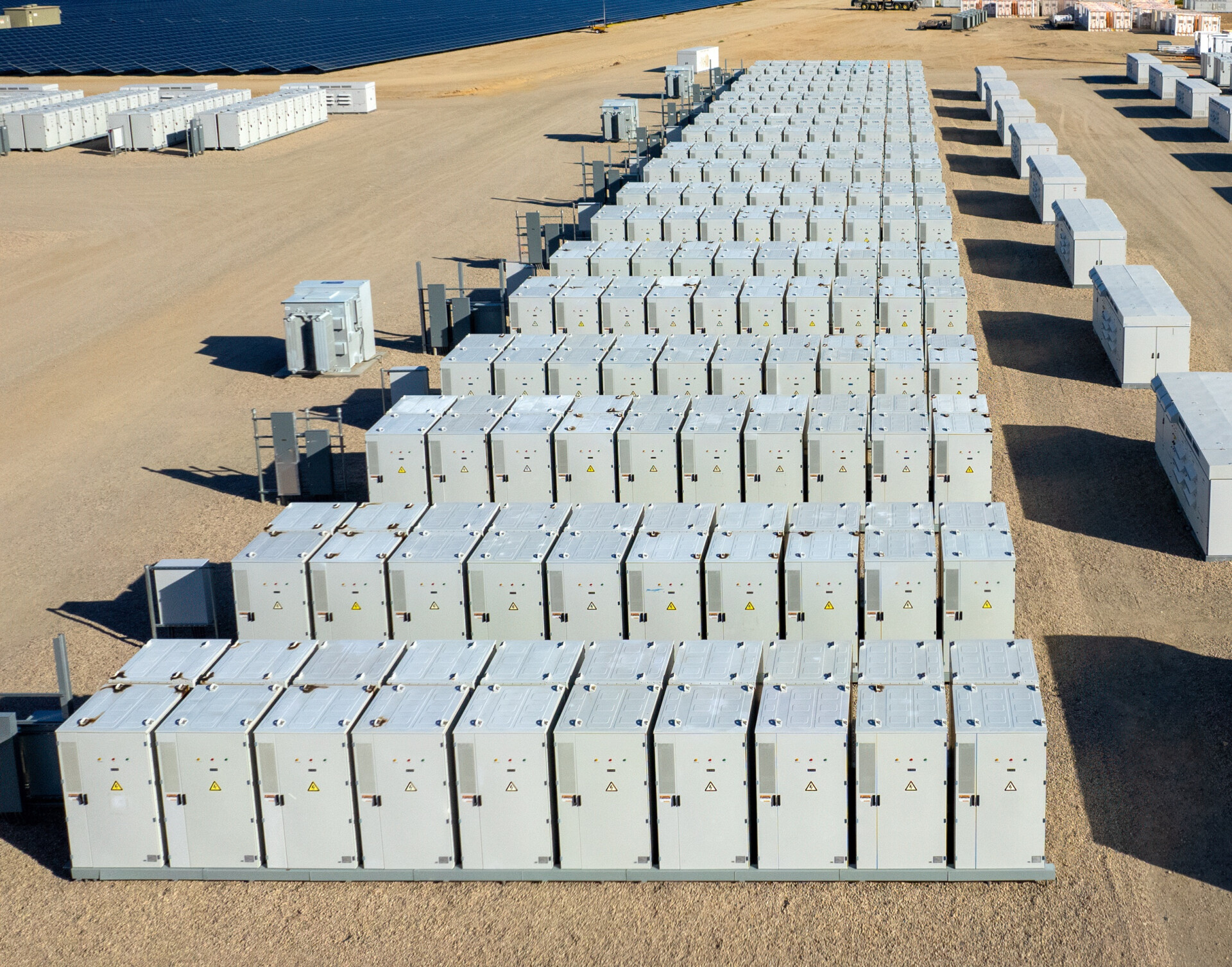 Aerial view of industrial battery units storing electricity in the desert. In the distance are solar panels and mountains.